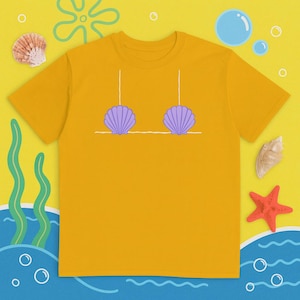 May include: Mustard yellow t-shirt featuring a cartoon illustration of a seashell bra in purple. The design includes white lines and is set against a beach-themed background with seashells, bubbles, and a starfish.