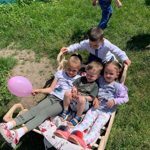 May include: A group of children are sitting in a wooden sled on a sunny day. One child holds a pink balloon. The sled is on a grassy area. Other children are standing nearby.