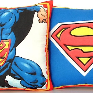 May include: Two square pillows with a Superman theme. One pillow features a comic book illustration of Superman in action. The other pillow has the Superman logo, a red and yellow emblem on a blue background. Both pillows have red and yellow borders.