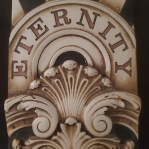 Könnte beinhalten: Rechteckige, cremefarbene Zierplatte mit dem Wort "ETERNITY" in braunen Buchstaben. Die Platte weist ein verziertes Design mit Schnörkeln und einem fächerförmigen Element auf. Schwarz-weiß karierte Akzente rahmen die Oberseite ein.