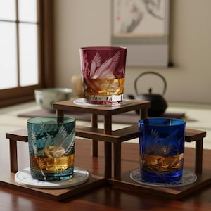 Peut inclure: Trois verres à boire colorés, gravés d'un motif de grue, remplis d'un liquide doré et de glaçons. Les verres sont présentés sur de petits supports en bois avec des dessous de verre, dans un style japonais.