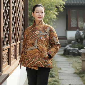 May include: A quilted jacket featuring a mandarin collar and a detailed crane and cloud design in gold, brown, and red hues. The jacket has a wrap-style closure and is worn with black trousers. The design of the jacket is inspired by traditional Asian art.