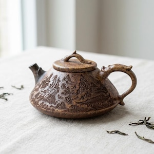 Peut inclure: Une théière en céramique marron avec un motif détaillé de paysage de montagne. La théière a une poignée incurvée et un couvercle avec une petite poignée en forme de branche. Le bec est petit et incliné. La théière est posée sur une surface blanche.