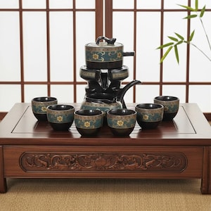 Può includere: Un tavolo da tè in legno scuro con un design floreale intagliato contiene un set da tè nero. Il set include una teiera, uno scaldavivande e sei piccole tazze da tè. Le tazze e la teiera hanno un motivo floreale verde acqua e oro. Un vaso con bambù è sullo sfondo.