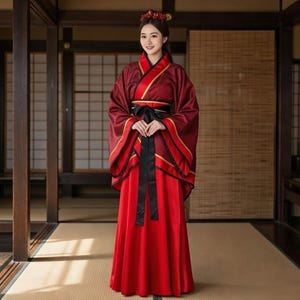 Peut inclure: Une femme portant une robe Hanfu traditionnelle rouge et bordeaux. La robe présente un col montant, des manches longues et une ceinture noire. Des bordures dorées ornent les manches et l'encolure. La femme porte un serre-tête floral.