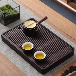 Puede incluir: Una bandeja de té de madera marrón oscuro con un diseño de listones, que contiene una tetera negra con tapa dorada y asa de madera, y dos pequeñas tazas de té llenas de té. También hay un pequeño terrario y una bandeja de bambú con hojas de té.