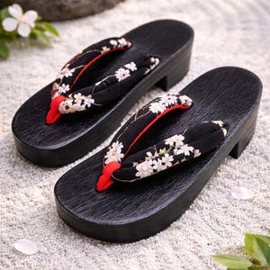 Puede incluir: Un par de sandalias geta japonesas tradicionales. Las plataformas de madera negra presentan correas de tela con un estampado floral en blanco y rojo. Las sandalias se exhiben sobre una superficie clara y texturizada.