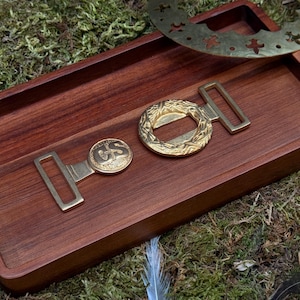 May include: Close-up of antique brass belt buckles and a decorative ring on a wooden tray. The buckles have intricate designs and are set against a backdrop of moss and a single feather. The overall aesthetic is vintage and rustic.