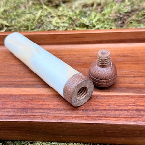 May include: A wooden pipe with a light-colored body and dark wood ends, along with a small, round wooden piece. The items are displayed on a reddish-brown wooden tray, with a mossy green background.