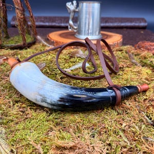 May include: A horn-shaped container with a leather strap, featuring a gradient of white, black, and brown. The container rests on a bed of green moss, with a small metal mug and wooden slice in the background. The horn is likely used for carrying liquids.