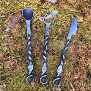 May include: Hand-forged metal flatware set including a spoon, fork, and knife. Each piece has a twisted handle with a decorative design near the base and a loop at the end. The set is displayed on a bed of green moss.