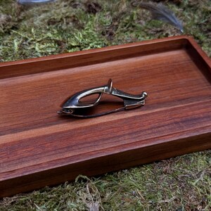 May include: A silver-toned metal brooch in the shape of a fish, resting on a dark wooden tray. The brooch has a pin closure on the back. The tray sits on a bed of green moss, with a few feathers scattered around.