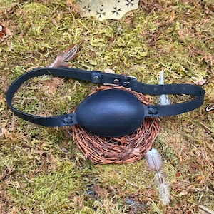 May include: A black leather eye patch with an adjustable strap and buckle. The eye patch is oval-shaped and sits atop a small woven basket. The background features green moss and a few feathers.