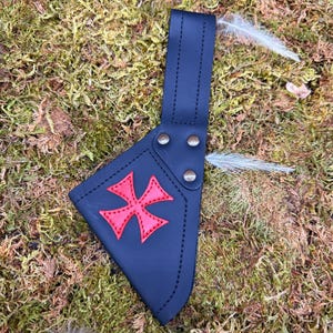 May include: A navy blue leather axe sheath with a red cross design. The sheath has a strap with rivets and is stitched with white thread. The background is a bed of green moss with a few feathers.