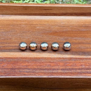 May include: Five small, shiny, silver-colored metal spheres are arranged in a row on a wooden tray. The tray has a rich, dark brown color and is set against a backdrop of green moss.