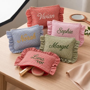 May include: Five rectangular zipped pouches with ruffled edges and gingham patterns in red, light blue, pink, green, and red. Each pouch features embroidered names: Vivian, Norah, Sophie, Margot, and Luna. One open red pouch reveals a gold lipstick and compact.