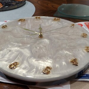 Könnte beinhalten: Eine runde Wanduhr mit einem weißen und goldenen Harz-Zifferblatt. Die Uhr hat goldfarbene Ziffern und Zeiger. Die Uhr hat einen Durchmesser von etwa 25 cm.