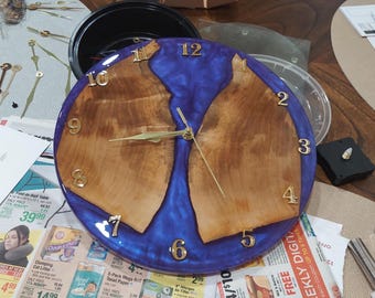 Puede incluir: Un reloj de madera hecho a mano con un centro de resina azul. La esfera del reloj presenta números y manecillas doradas. Las secciones de madera son de color marrón claro y el reloj tiene unos 25 cm de diámetro.