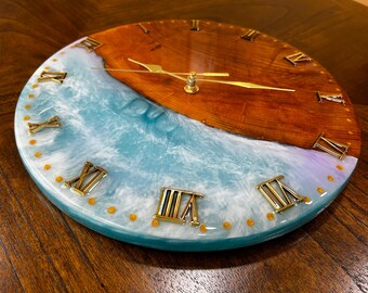 Puede incluir: Reloj de pared redondo de madera con un diseño único. La mitad superior es de madera natural, mientras que la mitad inferior presenta un diseño de olas de resina azul y blanca. Números romanos y manecillas doradas. Diámetro aproximado de 30 cm.
