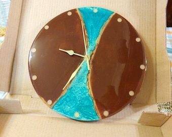 Puede incluir: Un reloj de pared redondo con un diseño marrón y turquesa. La esfera del reloj está dividida en secciones marrones y turquesas, con manecillas doradas y marcadores de hora de puntos blancos. El reloj está en una caja de cartón.