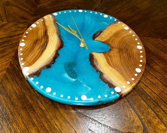 Puede incluir: Reloj de madera redondo con centro de resina turquesa y bordes de madera natural. El reloj tiene manecillas doradas y detalles de puntos blancos alrededor del perímetro. El reloj está sobre una superficie de madera oscura.