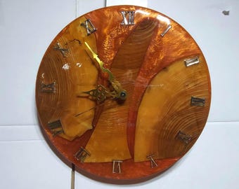 May include: Round wooden clock with a resin finish. The clock face features natural wood grain patterns and embedded Roman numerals. The clock hands are gold-colored. The resin has an orange hue.