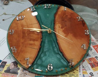 Puede incluir: Un reloj de madera redondo con un centro de resina turquesa y bordes de madera natural. Los números plateados marcan las horas, y las manecillas doradas indican la hora. El diseño del reloj combina elementos naturales con una estética moderna.