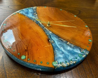 Puede incluir: Reloj de madera redondo con diseño de río de resina azul. La esfera del reloj presenta tonos de madera natural, manecillas doradas y detalles turquesa en el borde. Un reloj de pared decorativo único.