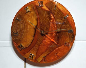 May include: A round wall clock with a wood grain design and orange resin. The clock face features Roman numerals and gold-colored clock hands, with an orange second hand. The clock's design incorporates natural wood elements.