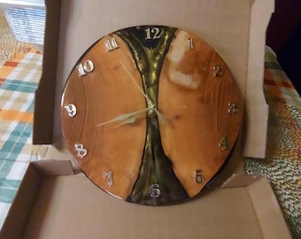 May include: A round wooden wall clock with a natural wood grain finish. The clock face features silver-colored numerals and hands, with a dark green and gold resin center. The clock is displayed inside a cardboard box.