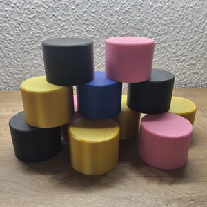 Könnte beinhalten: Eine Sammlung zylindrischer Objekte in verschiedenen Farben, darunter Schwarz, Rosa, Gelb und Blau. Die Objekte sind auf einer Holzoberfläche gestapelt, wodurch eine visuell interessante Anordnung entsteht. Die Zylinder haben eine einheitliche Höhe und einen einheitlichen Durchmesser.