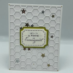 May include: A white greeting card with a honeycomb pattern background and gold star embellishments. The card features a decorative label with the words "A Note of Encouragement" in elegant script. The card has a gold border.