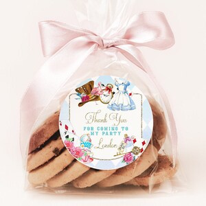 May include: A clear cellophane bag filled with cookies tied with a pink ribbon. A round sticker with a whimsical Alice in Wonderland theme is attached to the bag. The sticker features Alice, the Mad Hatter, and a pocket watch. The text on the sticker reads "Thank You for coming to my party London."