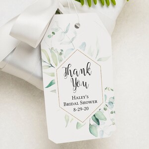 May include: A white gift box with a white satin ribbon and a green leafy sprig. A white gift tag with a gold hexagon frame and green watercolor leaves reads "Thank You Haley's Bridal Shower 8-29-20".