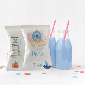 May include: Two white bags with a Cinderella carriage design and the text "Lulu's is Two" and a glass slipper. The bags are filled with snacks and are sitting on a white surface with blue, pink, and gold confetti.