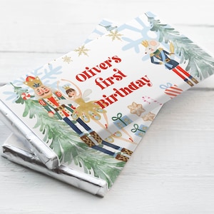 May include: Two wrapped chocolate bars with a festive design. The wrappers feature illustrations of a nutcracker, a ballerina, and a mouse king, along with the text "Oliver's first Birthday". The design includes snowflakes, gifts, and stars.