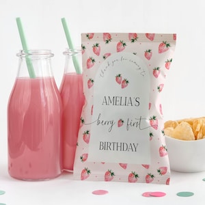 May include: Pink and white favor bag with a strawberry pattern. The bag has a white label with the text "Amelia's berry first birthday" printed on it.
