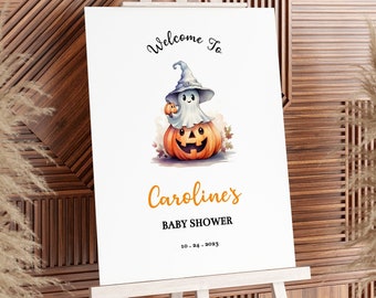 May include: A white welcome sign with a cartoon ghost wearing a witch hat and holding a small pumpkin. The ghost is sitting on top of a large orange pumpkin with a jack-o-lantern face. The sign reads "Welcome to Caroline's Baby Shower 10.04.2023".