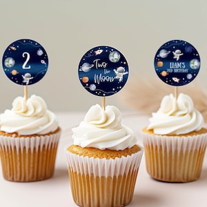May include: Three cupcakes decorated with blue and white space-themed toppers. One topper says "Two the Moon", another says "Liam's 2nd Birthday", and the last one has the number "2" on it.