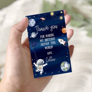 Puede incluir: Una tarjeta de agradecimiento azul con temática espacial. Presenta un astronauta, planetas, estrellas y un cohete. El texto dice "¡Gracias por hacer que mi cumpleaños sea fuera de este mundo! Con cariño, Collin."