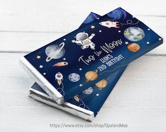 May include: Blue and silver chocolate bar wrapper with a space theme. It features an astronaut, planets, stars, and rockets. The text reads "Two the Moon Liam's 2nd Birthday".
