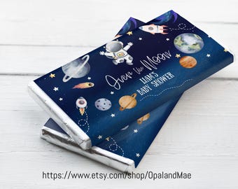 Könnte beinhalten: Blaue und weiße Schokoladentafel-Verpackung mit einem Astronauten, Planeten, Sternen und Raketen. Der Text auf der Verpackung lautet "Over the Moon Liam's Baby Shower".