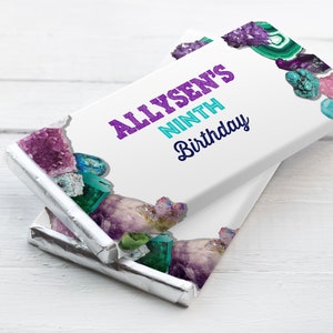 Puede incluir: Dos barras de chocolate envueltas en papel de aluminio plateado con una etiqueta blanca. La etiqueta tiene un diseño morado y turquesa con el texto "Allysen's Ninth Birthday".
