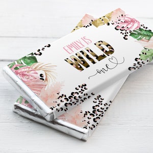 Könnte beinhalten: Ein weißer Schokoladenriegel-Wrapper mit einem tropischen Blattdesign in Pink, Gold und Grün. Der Text "Emily is Wild one" ist auf den Wrapper gedruckt.