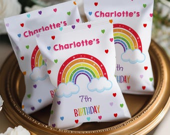 May include: Three white party favor bags with a rainbow and cloud design, scattered hearts, and the text "Charlotte's" and "7th Birthday". The bags are on a gold-colored tray.