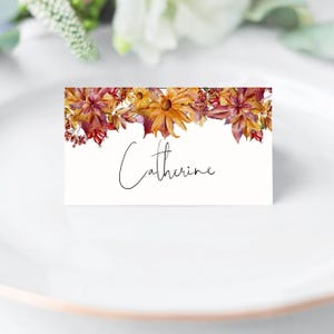 Könnte beinhalten: Eine weiße Tischkarte mit einem Aquarell-Design aus Herbstblättern und Blumen in Rot-, Orange- und Gelbtönen. Der Text "Catherine" ist in schwarzer Kursivschrift geschrieben.