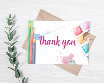 May include: A white thank you card with watercolor illustrations of paintbrushes, paint, and colored pencils. The text "thank you" is written in pink.