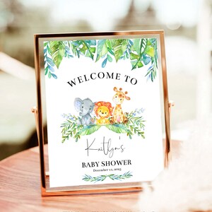 May include: A welcome sign for a baby shower with a jungle theme. The sign features a green leafy border with a giraffe, lion, and elephant. The text reads "Welcome to Kaitlyn's Baby Shower December 11, 2023".