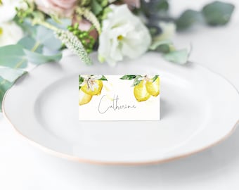 May include: A white place card featuring a watercolour lemon design and the name "Catherine" in script. The card sits on a white plate, with a blurred floral arrangement in the background.