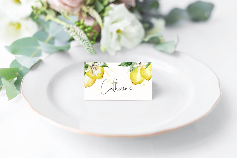 May include: A white place card with a watercolor lemon design and the name "Catherine" in script. The card is on a white plate, with a blurred floral arrangement in the background.